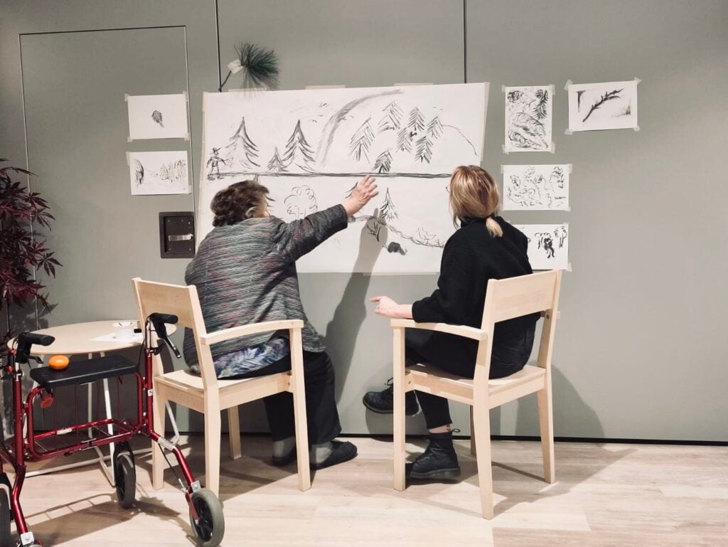 Iäkäs henkilö sekä nuorempi henkilö seinälle ripustettujen hiilipiirrosten edessä.