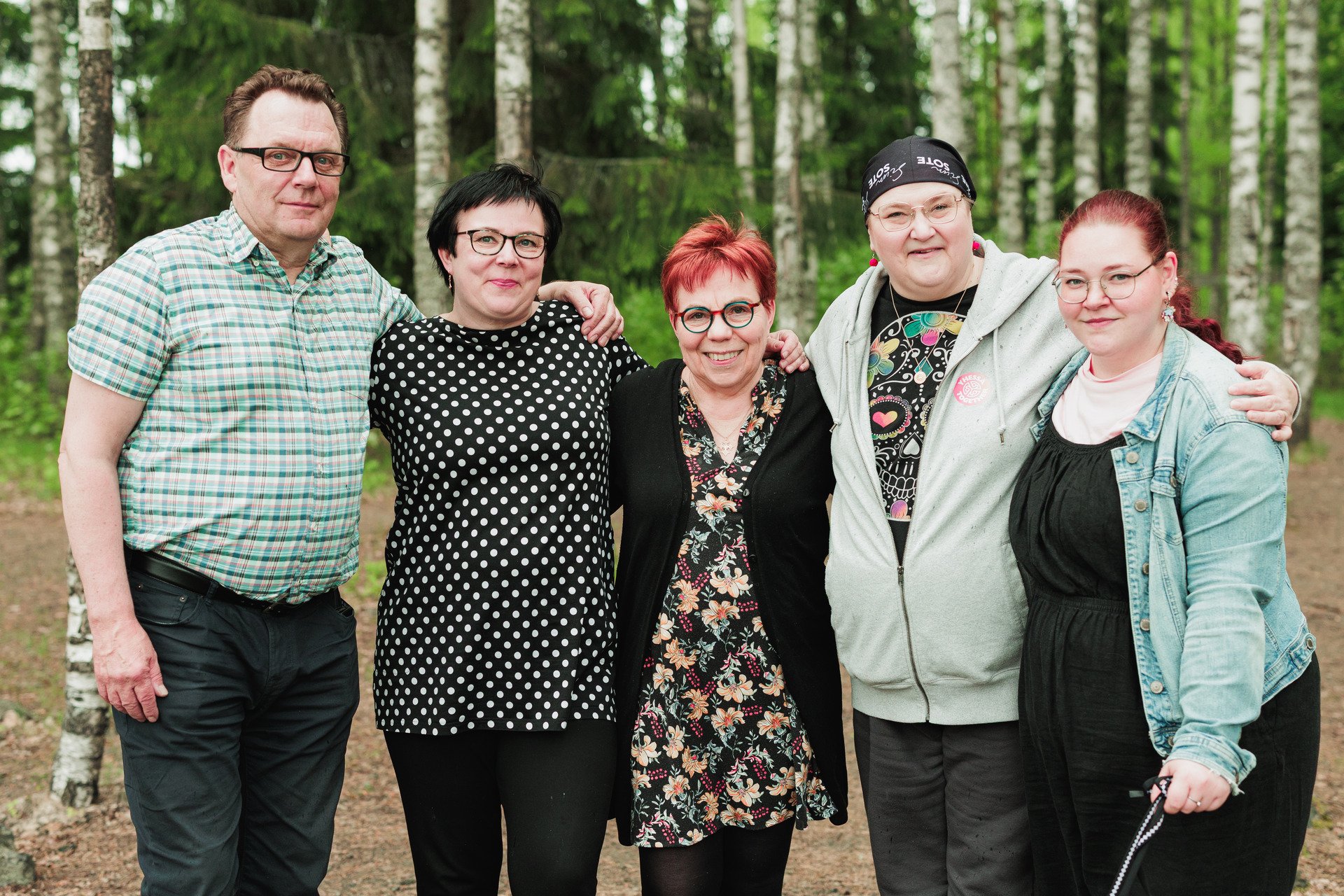 Viisi aikuista on rivissä, lähellä toisiaan, kädet toistensa hartioilla.