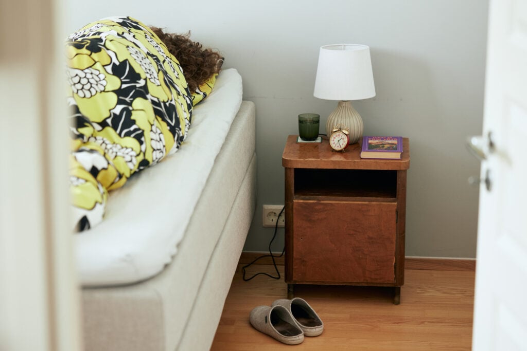 Kuvassa näkyy peiton alta nukkujan hiukset, vieressä on yöpöytä ja sängyn vieressä tohvelit.