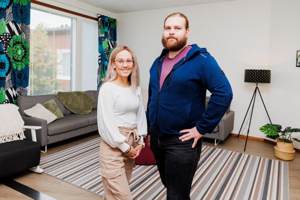 Kaksi henkilöä seisovat olohuonetilassa ja hymyilevät kameralle.