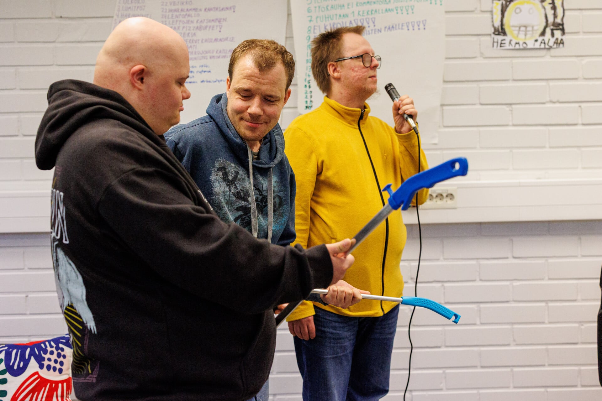 Kolme henkilöä, joista yksi laulamassa mikkiin ja kaksi pitävät rikkaharjoja käsissään.